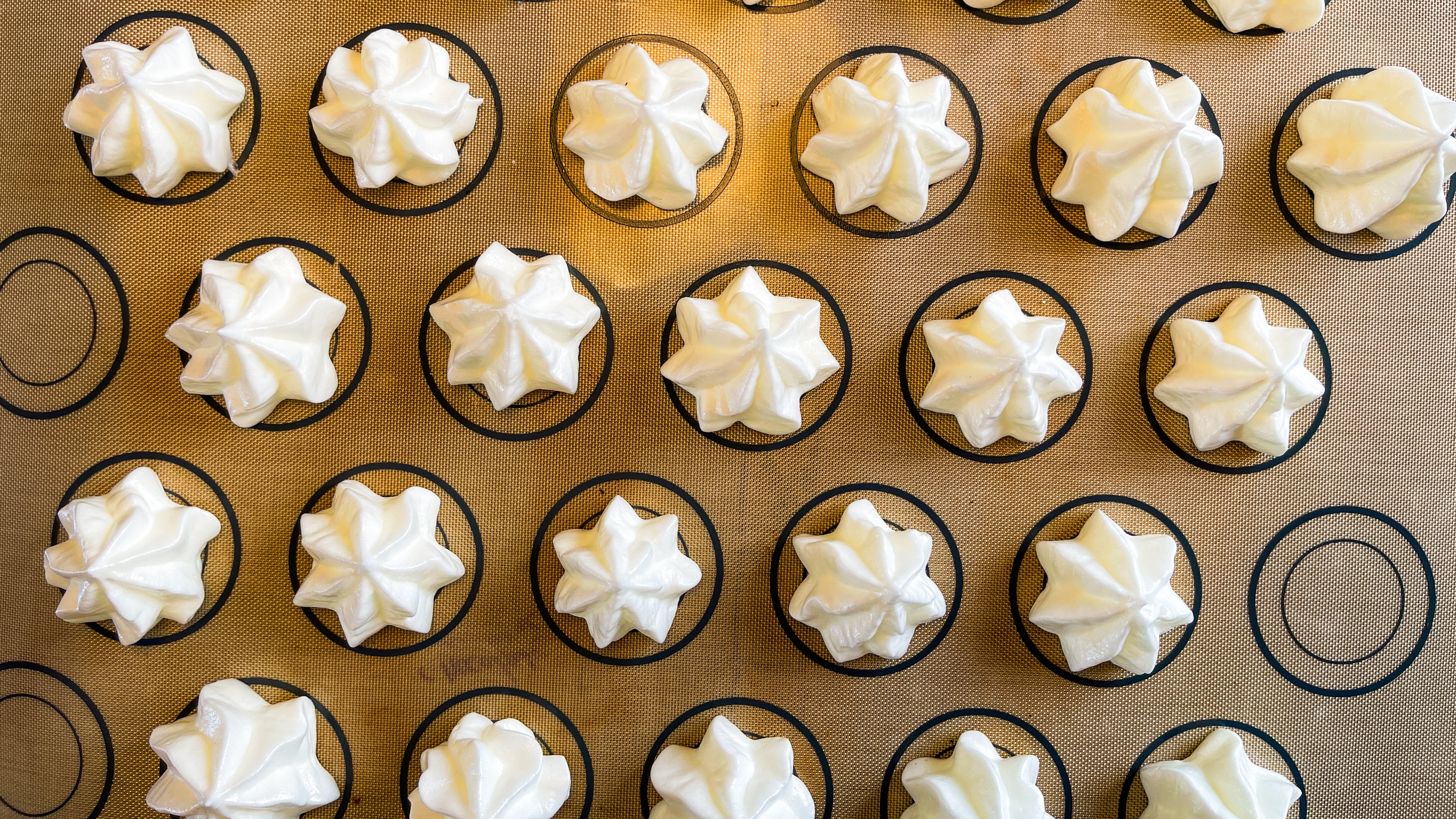 Rows of white meringue cookies are evenly spaced on a brown baking mat marked with black circles, viewed from above.