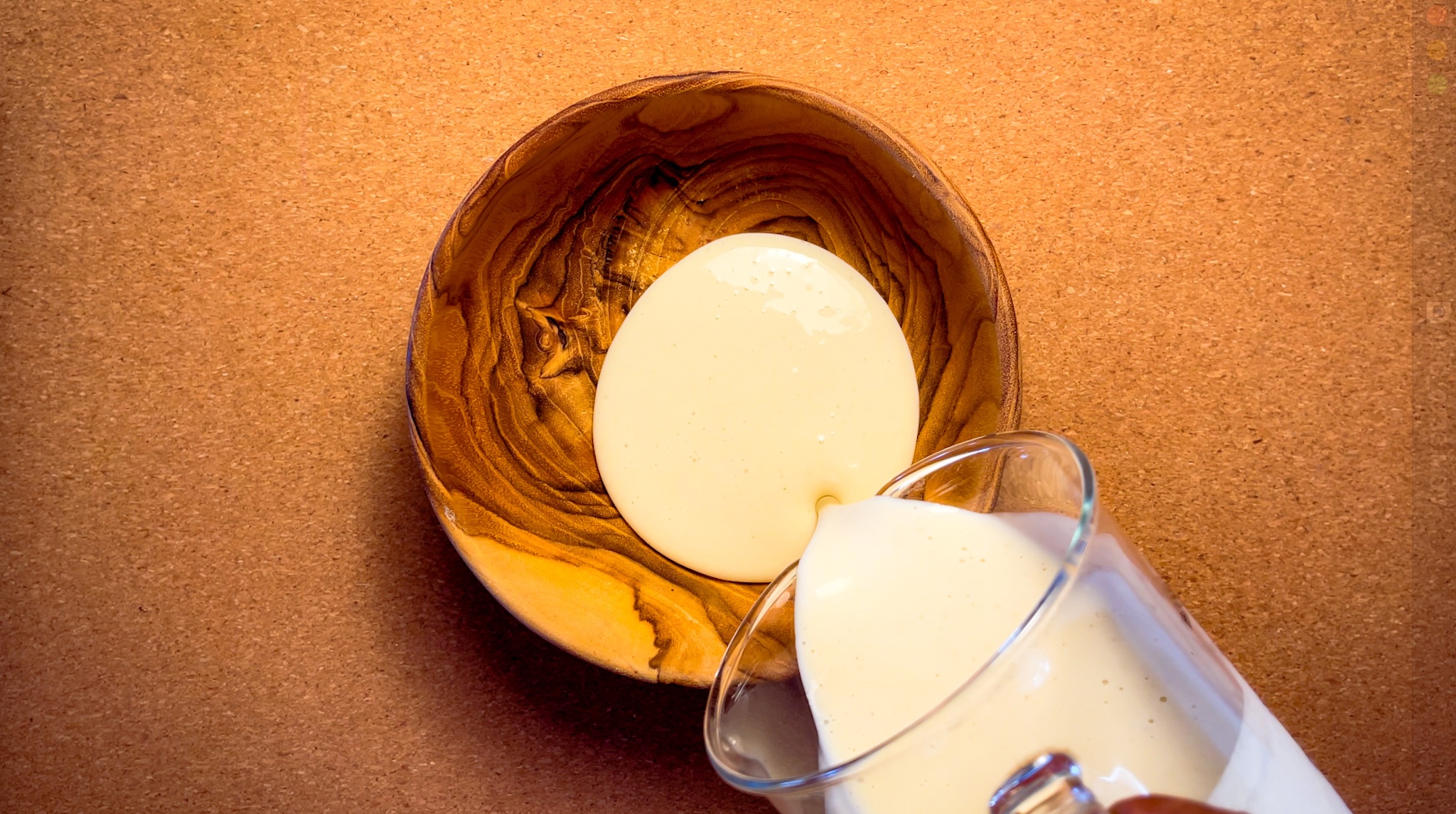 A glass of creamy liquid is being poured into a small wooden bowl on a cork surface.