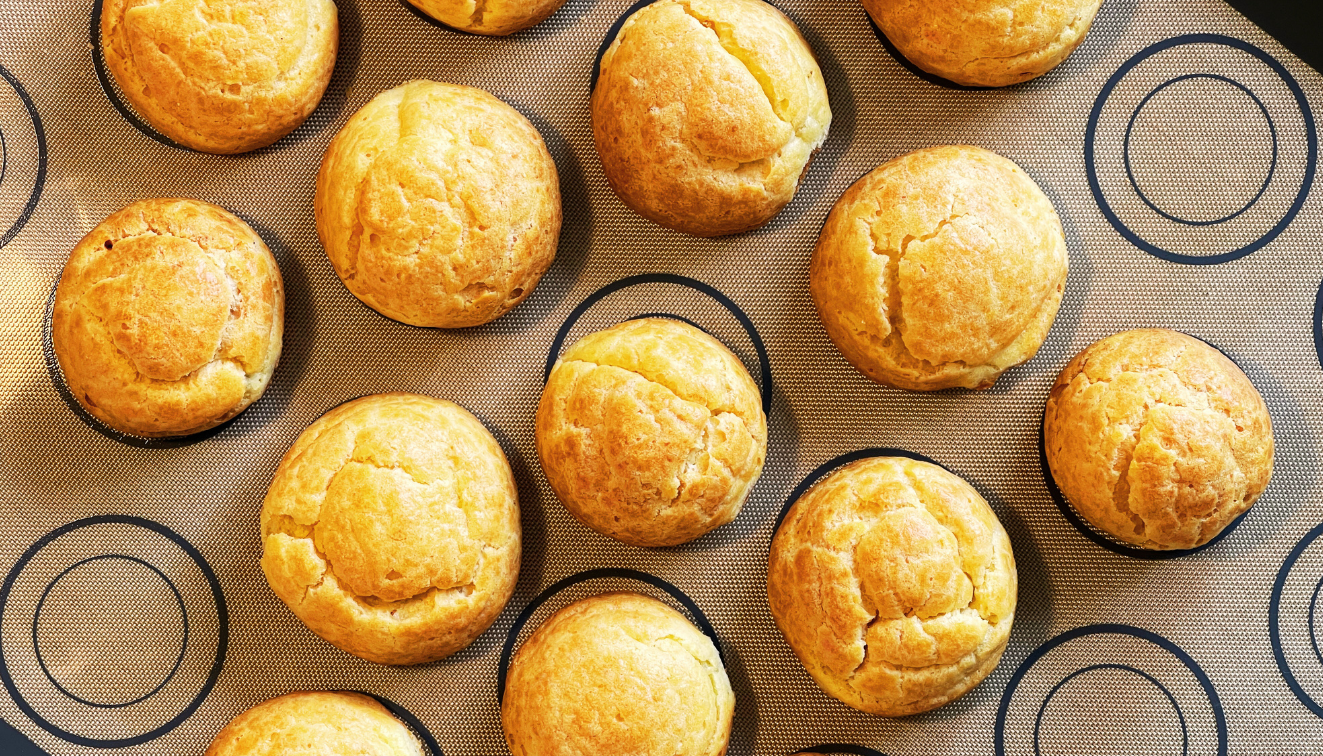 A top-down view of golden-brown muffins arranged on a baking mat with a pattern of black concentric circles. The muffins are evenly spaced and have a slightly cracked surface.