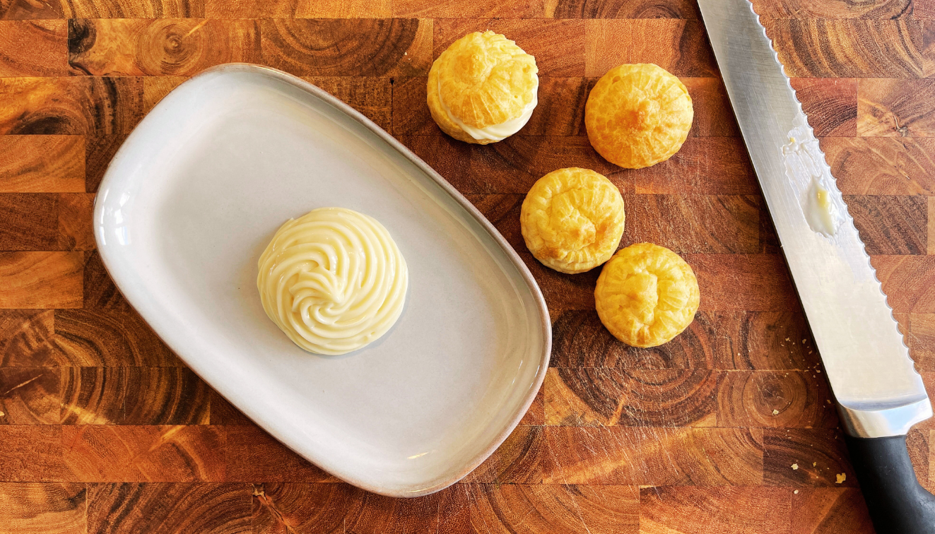 A swirl of piped cream sits on a white oval plate next to four small golden pastry puffs and a serrated knife on a wooden cutting board.