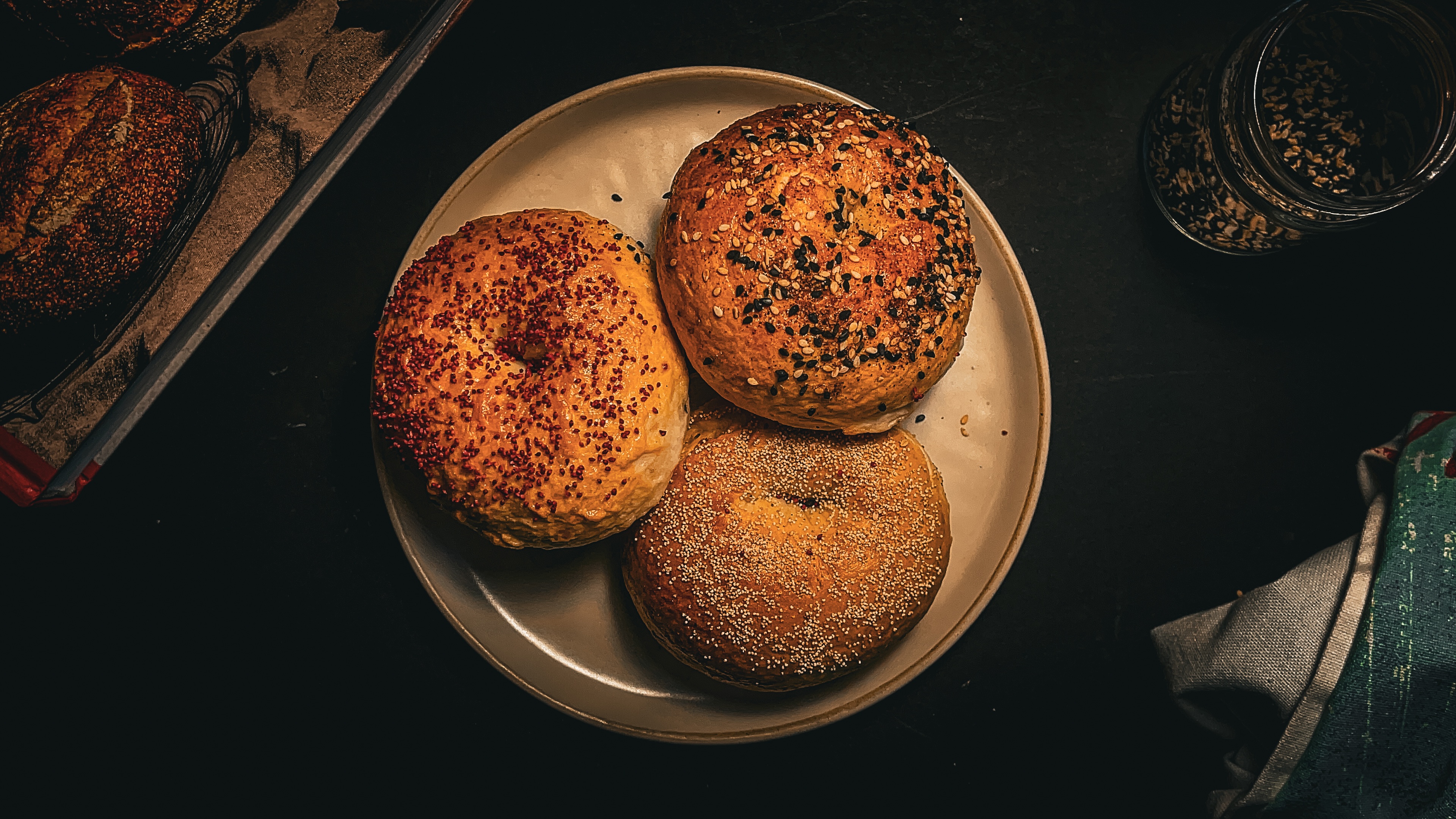 A plate with three bagels topped with different seeds sits on a dark surface; nearby are a jar with more seeds and part of a towel.