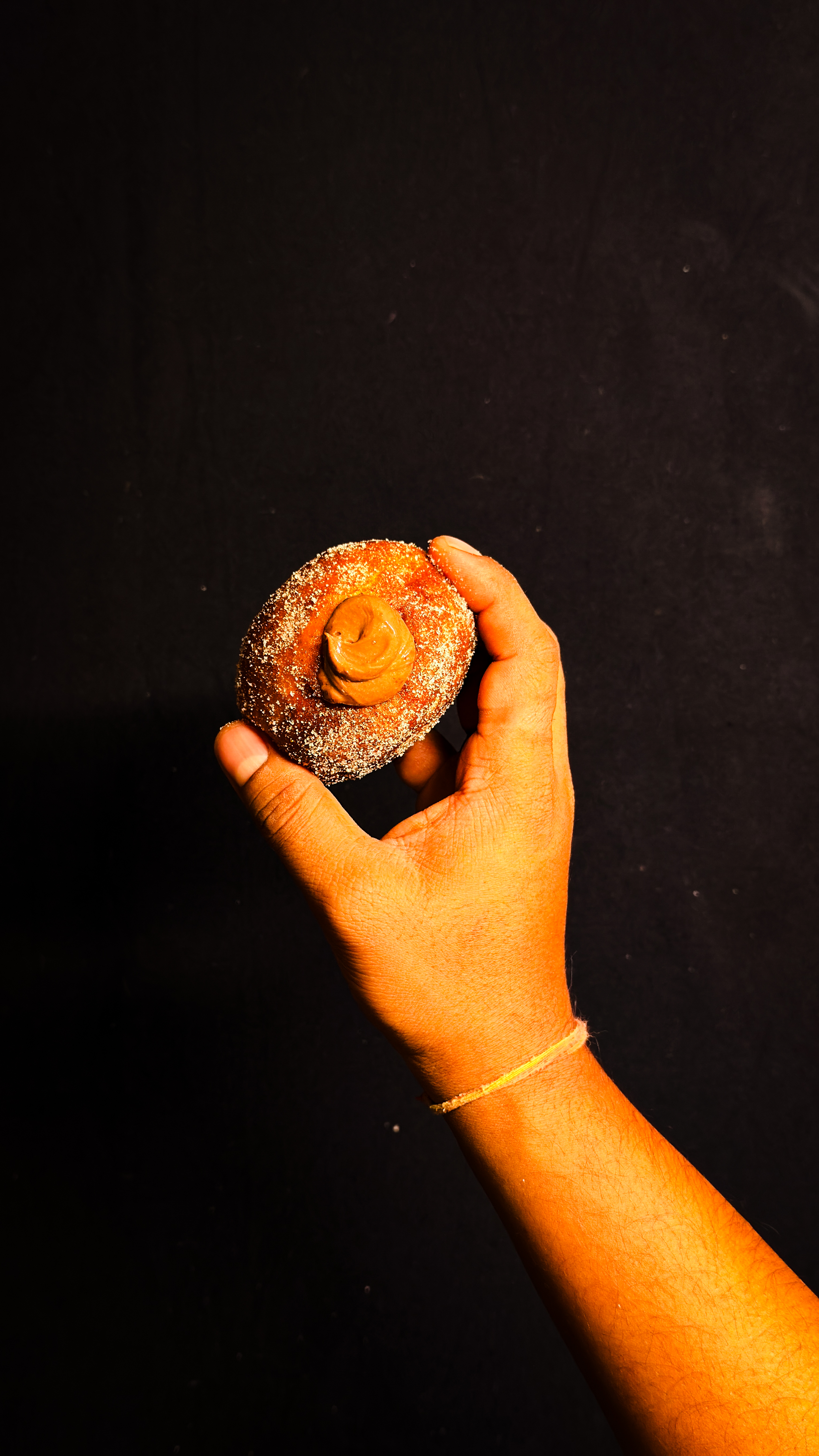 A hand holding a sugar-coated round pastry with a swirl of caramel filling on top, against a plain dark background.