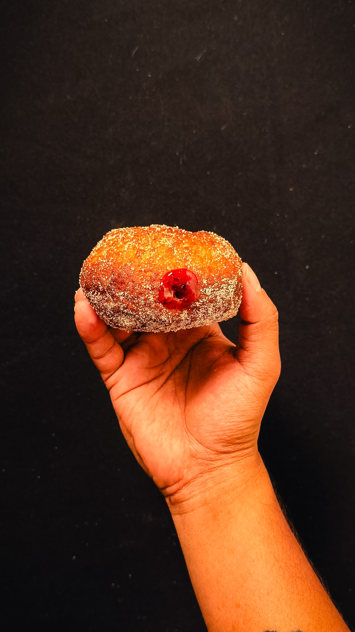 A hand holding a sugar-coated jelly-filled donut with red jelly oozing from the center against a plain dark background.