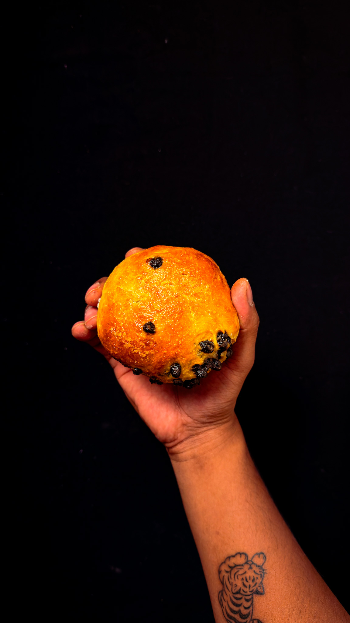 A hand with a tiger tattoo on the wrist holds a golden-brown, round bread roll with visible blueberries against a black background.